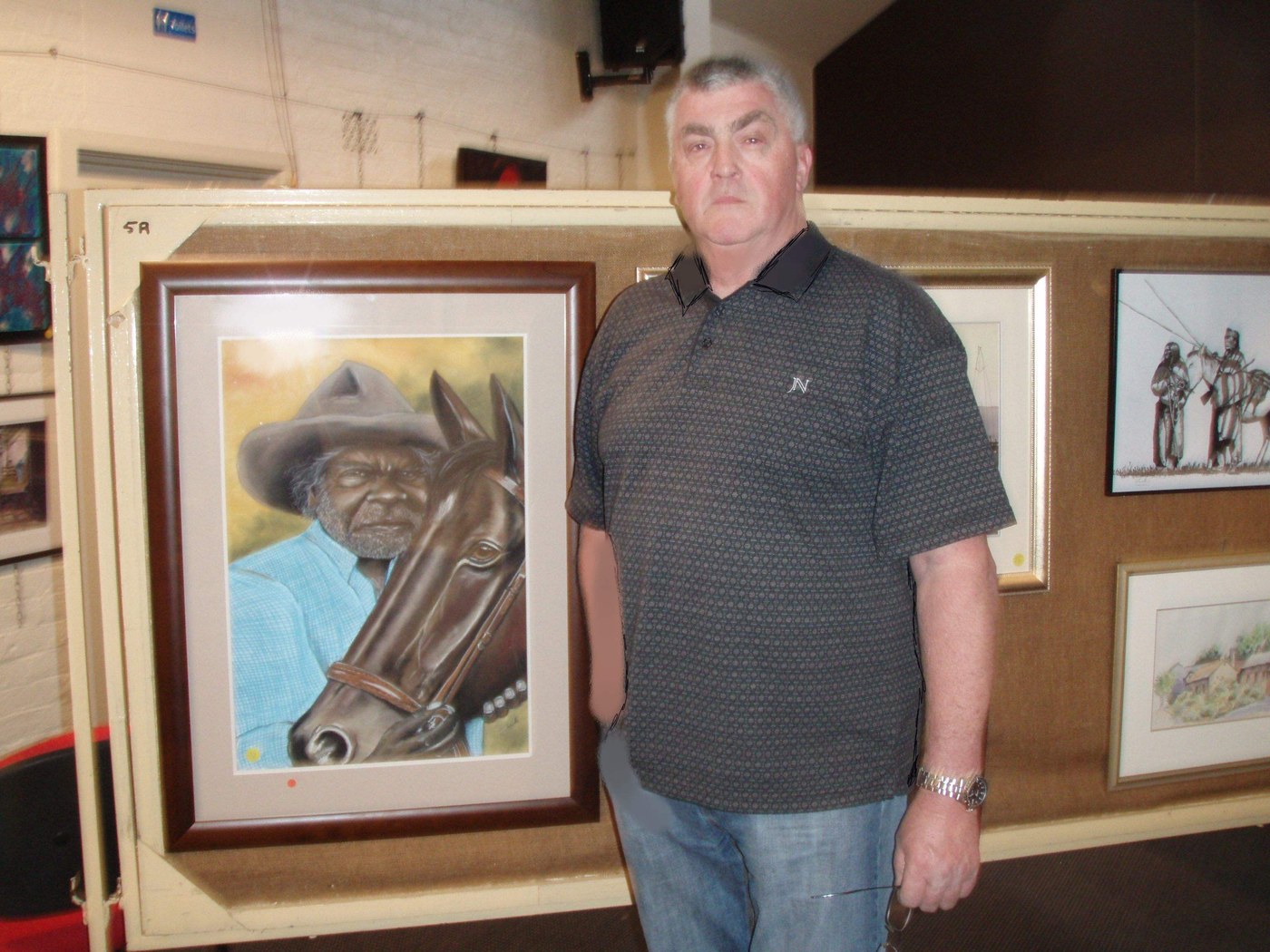 Colin Bridle standing beside his framed artwork at an exhibition